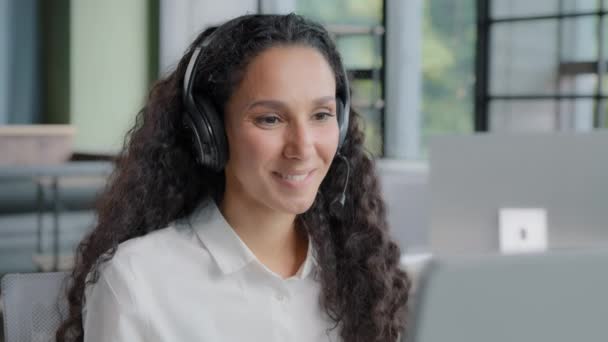 Young positive woman in headphones communicates via video call call center operator talks to customer on online conference via webcam on computer using headset distance office chat e-learning concept - 映像、動画