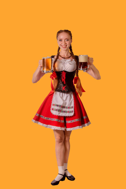 sexy oktoberfest waitress dressed in traditional bavarian dress serving two big mugs of beer isolated on yellow background - Foto, immagini