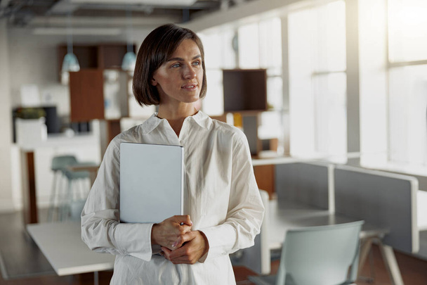Ελκυστική γυναίκα των ευρωπαϊκών επιχειρήσεων με φορητό υπολογιστή στέκεται στο σύγχρονο γραφείο και κοιτάζοντας πλευρά - Φωτογραφία, εικόνα