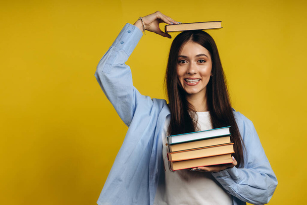 Portrait of student girl balancing books on head and hand while standing against yellow background - Photo, Image