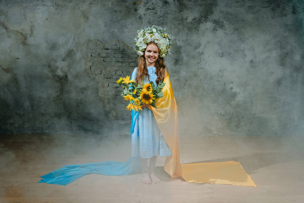 民族刺繍のドレスと花の花輪で小さな女の子は白い煙の中でロフト壁に対してウクライナの旗で立って - 写真・画像