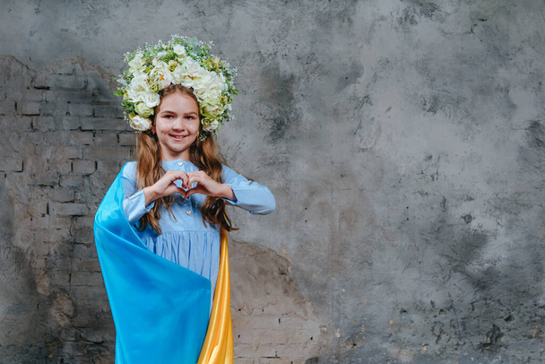 Little girl in flower wreath standing with Ukraininan flag against loft wall showing heart. Blank space for text. - Photo, Image