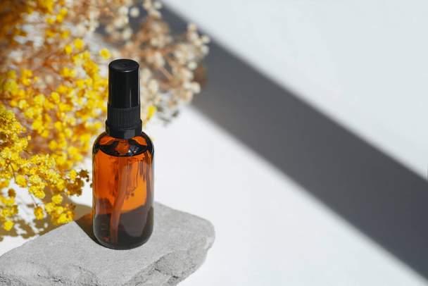 Amber pump bottle with serum, tonic or essential oil on grey concrete podium with yellow flowers. White background with daylight. Beauty concept for face and body care - Foto, Bild