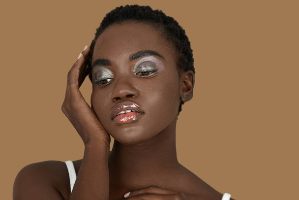 Closeup portrait of a serene young black woman with short Afro hair, light makeup and lipstick posing by herself inside a studio with a pecan background resting her palm of her hand on her face. - Photo, Image