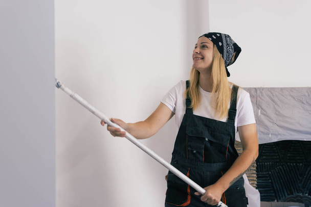 a happy girl with a black bandana on her head is dressed in a white T-shirt and a dark work overalls, paints a white wall with a roller. Gender equality, repair concept. High quality photo - Photo, Image
