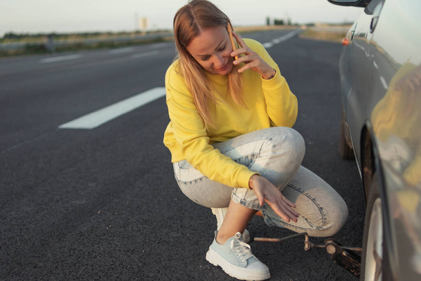 Дівчина в жовтому светрі й джинсах європейської зовнішності сидить біля заднього пробитого колеса в руках тримає ключ для коліс і говорить по телефону, як робити колесо. Фотографія високої якості - Фото, зображення