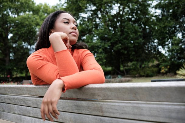 Young mixed race woman is sitting on a bench in a park and posing looking away. - Fotografie, Obrázek