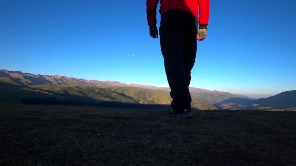 The guy goes to the edge of the hill and admires the view. In the distance there are high mountains and green hills covered with forest. Wide margins. The moon is visible in the blue sky. Sunrise - Materiał filmowy, wideo