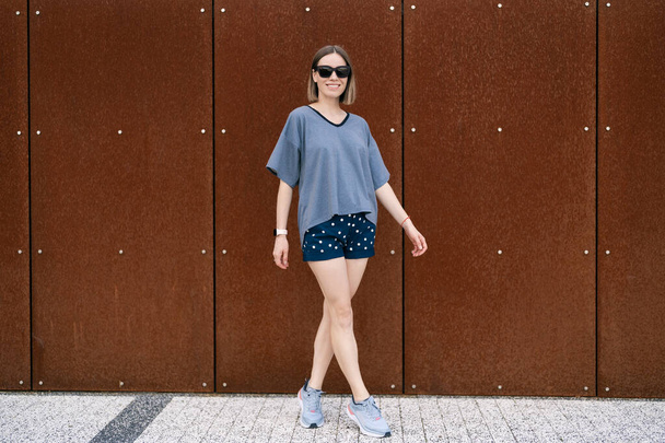 Young woman in casual t-shirt, shorts in sunglasses posing on rusty wall background. Urban style - Photo, Image