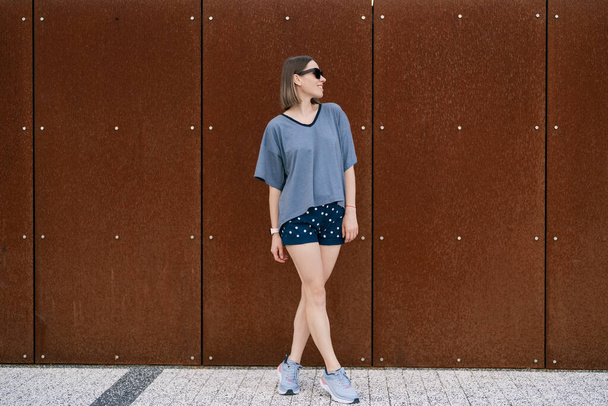 Молодая женщина в повседневной футболке, шорты в солнечных очках, позирующие на ржавом фоне стен. Городской стиль - Фото, изображение