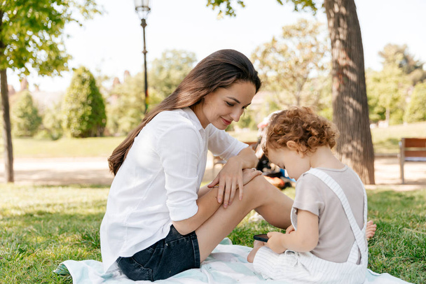 Pretty young woman with long dark hair sitting in the park on bright blanket with her little daughter and looking in smartphone. Woman spend time with daughter  - Foto, Imagem