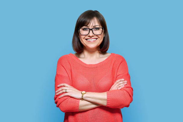 Attractive middle aged woman looking at camera on blue color background. Smiling confident 40s female with glasses, crossed arms in red casual clothes. Lifestyle, confidence, calmness, mature people - 写真・画像