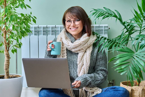 Mature woman in woolen clothes warming with cup of hot drink sitting near heating radiator, smiling female looking at camera using laptop at home. Heating season autumn winter lifestyle work freelance - Zdjęcie, obraz