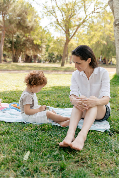 Full-length photo of young woman with dark hair wearing white shirt and shorts sitting in park with little daughter and having fun.  - Фото, зображення