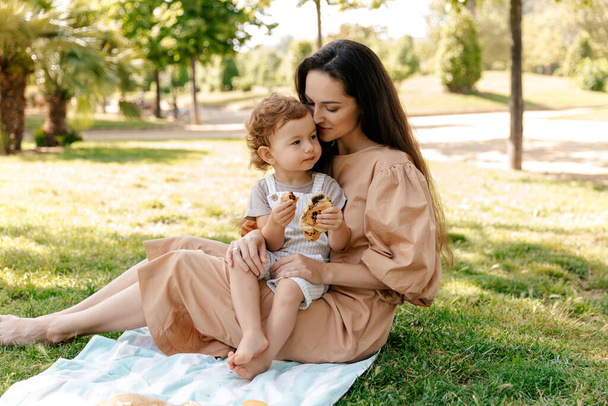 Close up outdoor photo of long-haired woman in summer dress holding her little daughter on hands. Charming little girl with curls spend day with mother in green park. - Fotoğraf, Görsel