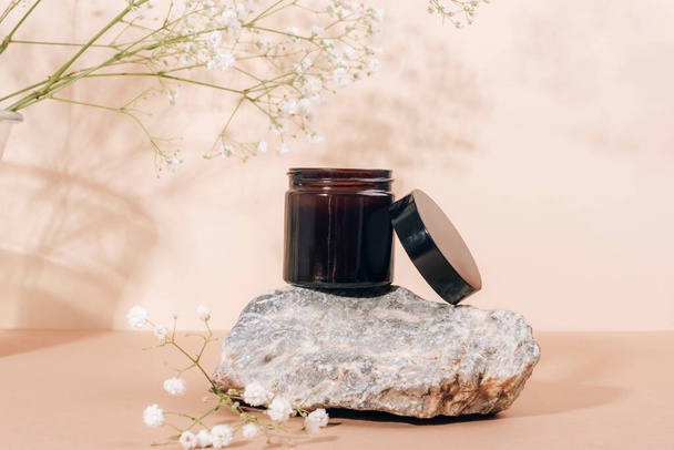 Cream in brown glass jar on natural stone with gypsophila flowers in sunlight. Neutral beige background, closeup. - 写真・画像
