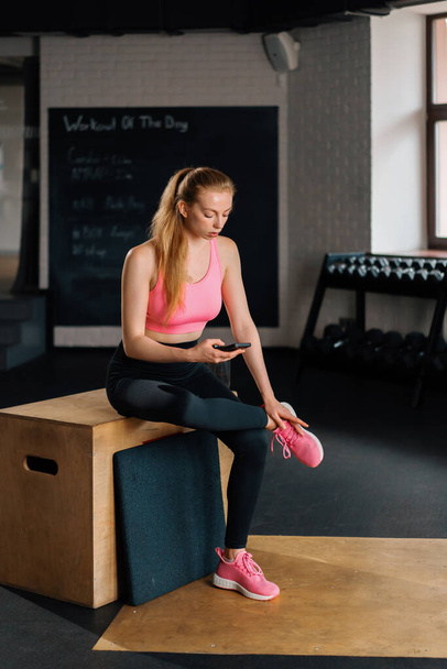 Athletic build girl uses the phone while resting after a grueling workout in t gym. - Фото, изображение