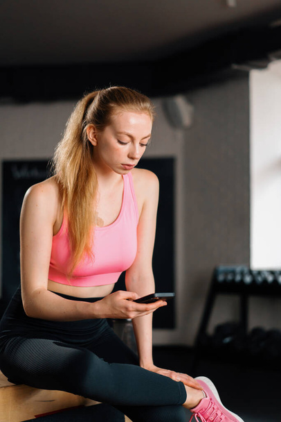 Спортивная телосложение девушка использует телефон во время отдыха после изнурительной тренировки в спортзале. - Фото, изображение