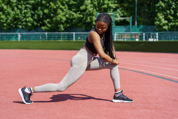 Афроамериканська леді у спортивному ласі насовує глиняні м'язи на спортивному майданчику біля буйних зелених дерев, що ростуть у міському парку. Чорна жінка з довгими плесками в сонячний літній день добре займається фізичними вправами. - Фото, зображення