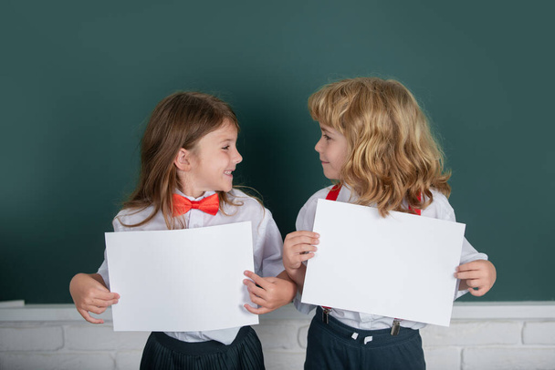 Two schoolkids holding white paper blank, poster with copy space. Schoolgirl and schoolboy in classroom at school. Friendship and classmate. Best school friends. Blank paper for children education - Fotó, kép