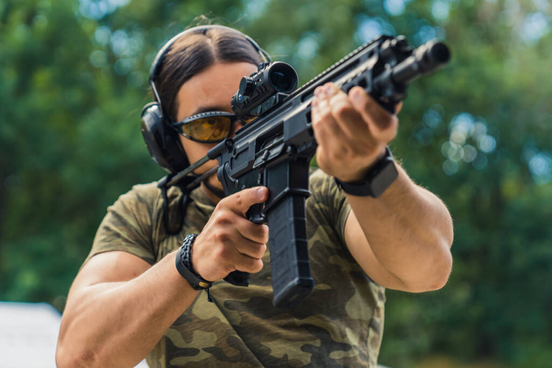 Military concept. Middle-aged European white black-haired man focused on aiming at target via rifle loupe. Medium closeup shot. Blurred trees in the background. High quality photo - Foto, imagen