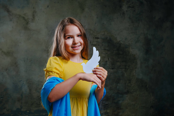 Niña con bandera ucraniana sosteniendo paloma de papel blanco como símbolo de oración y paz para Ucrania contra el fondo del loft oscuro. - Foto, Imagen