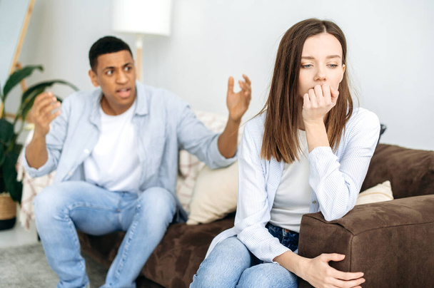 Mixed race couple sitting on a sofa in the living room, an angry man yells at a woman, feels angry, frustrated sad girl turned away from him. Misunderstanding in relationships, jealousy, quarrel - Photo, Image