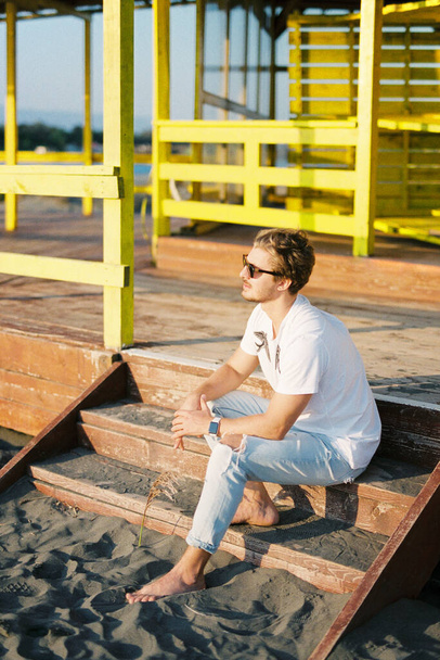 Young man in sunglasses sitting on the steps of a wooden building on the beach. High quality photo - Fotó, kép