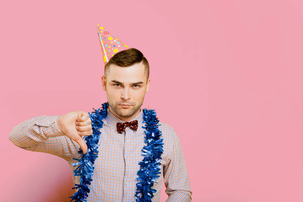 Young adult guy 20-25s wearing a party hat and New Year's tinsel, unhappy and angry showing a thumbs down or dislike gesture, frustrated with a displeased expression. - Photo, Image