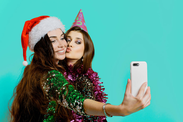 Young adult girls 20-25s in a party hat and santa claus cap and tinsel take a selfie or shoot a video on a smartphone for social networks on a mint isolated background with copy space - Фото, изображение