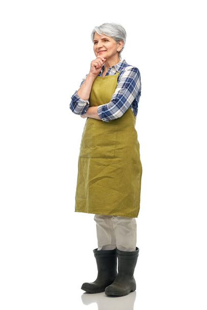 gardening, farming and old people concept - thinking senior woman in green garden apron and rubber boots over white background - Foto, afbeelding