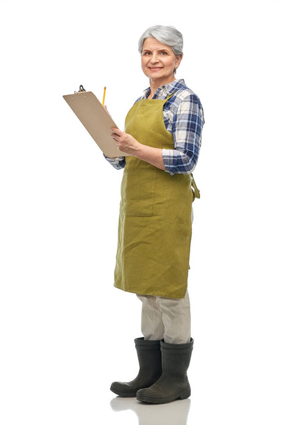 gardening, farming and old people concept - portrait of smiling senior woman in green garden apron and rubber boots with clipboard over white background - Φωτογραφία, εικόνα