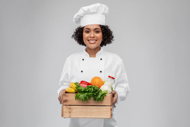 cooking, culinary and people concept - happy smiling female chef in toque holding food in wooden box over grey background - Фото, изображение