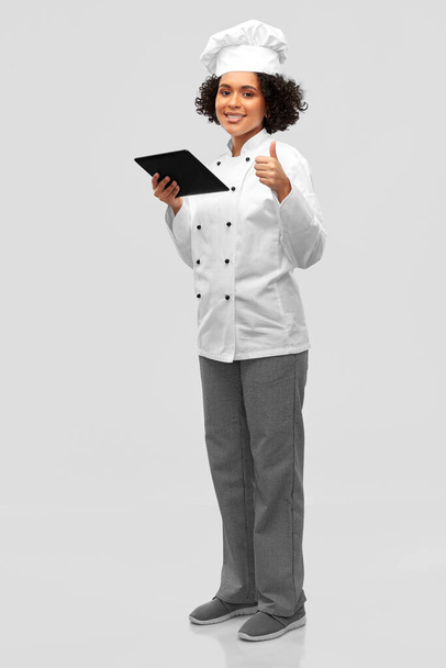 cooking, culinary and people concept - happy smiling female chef in toque and jacket with tablet pc computer showing thumbs up over grey background - Photo, Image