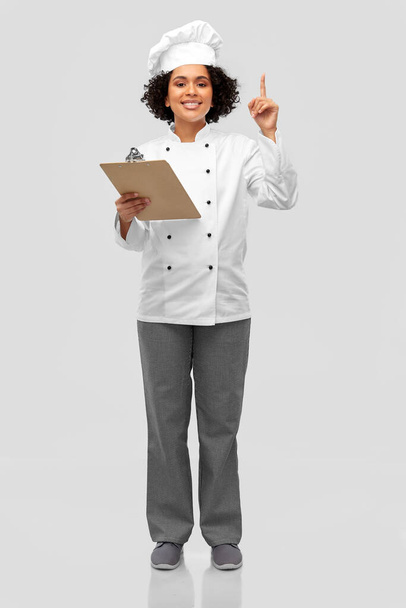 cooking, culinary and people concept - happy smiling female chef in toque and jacket with clipboard showing thumbs up over grey background - Photo, image