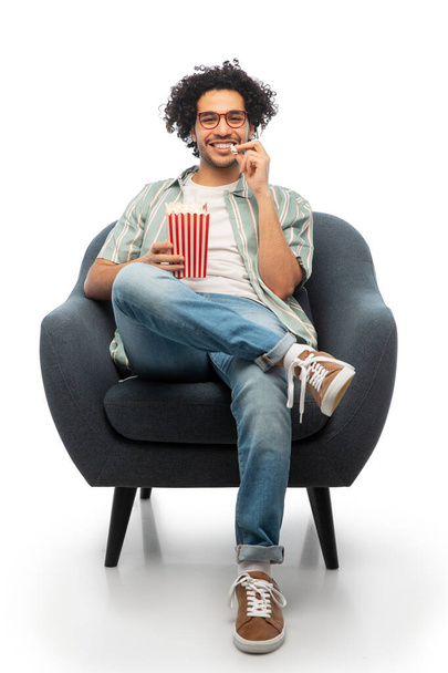 people and furniture concept - happy smiling young man in glasses with popcorn sitting in chair over white background - Fotó, kép