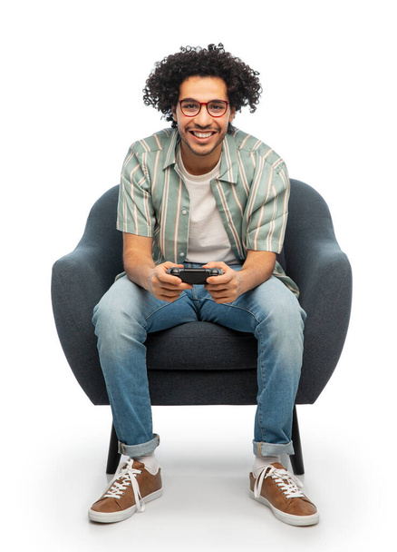 technology, people and leisure concept - happy smiling young man in glasses with gamepad sitting in chair and playing video game over white background - Photo, Image