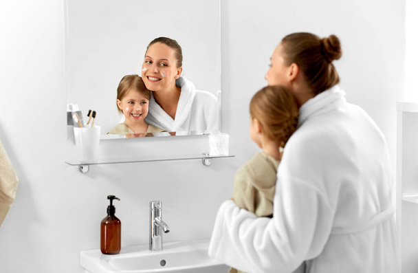 beauty, family and people concept - happy smiling mother and daughter with moisturizer on their faces looking to mirror in bathroom - Foto, Bild