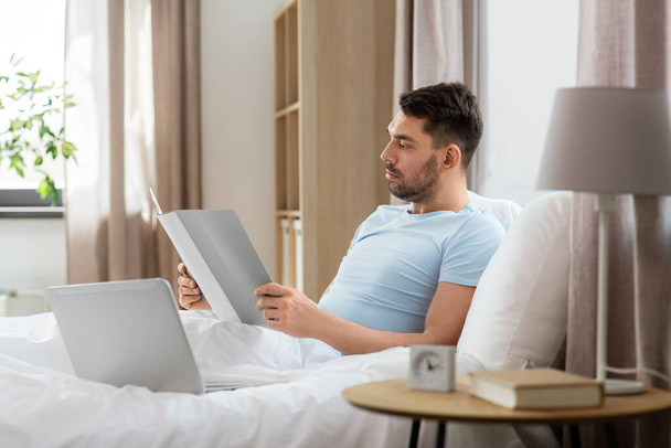 people, technology and remote job concept - man with laptop computer and folder working in bed at home bedroom - Φωτογραφία, εικόνα