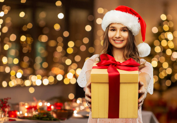christmas, holidays and people concept - happy smiling teenage girl in santa helper hat holding gift box over festive lights at home on background - Фото, изображение