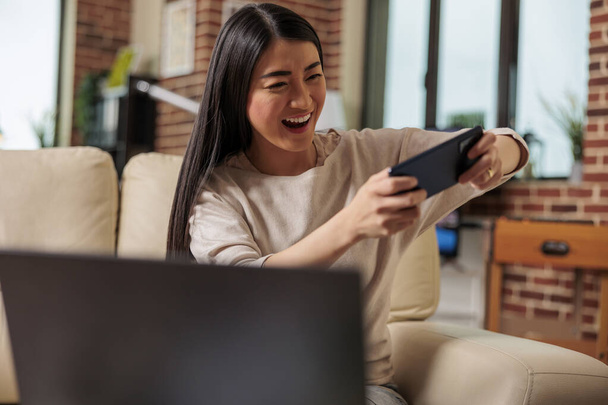 Autentyczna azjatycka kobieta grająca w mobilne gry wideo online na telefon wygrywające i szczęśliwe emocje. Internetowe gry wideo aplikacje do gier oglądanie filmów social internet komunikacji - Zdjęcie, obraz