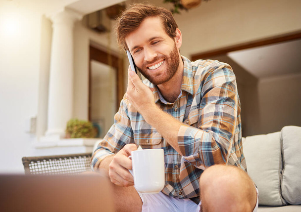Communication, coffee and man with phone on sofa to relax in living room. Success, internet and working from home with tea. Happy man sitting, relaxing with a laptop and on mobile call on the weekend. - 写真・画像