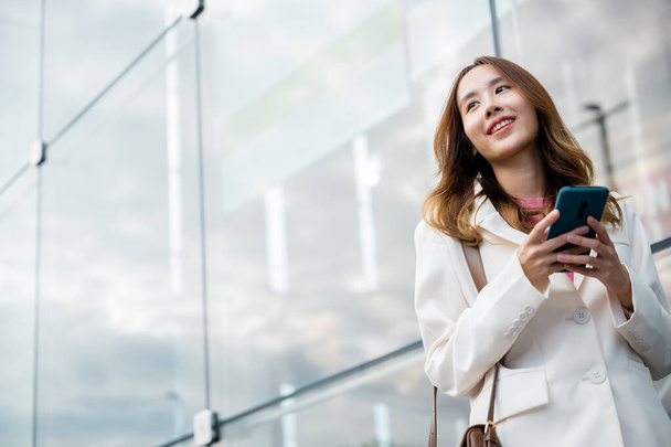 Asian businesswoman working with mobile phone standing against street front building near office, Portrait successful business woman smiling holding smartphone use application chat online in morning - Foto, imagen