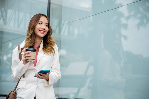 Young business woman smiling holding mobile phone with coffee take away going to work early in morning, Asian businesswoman with smartphone and cup coffee standing against street building near office - Photo, image
