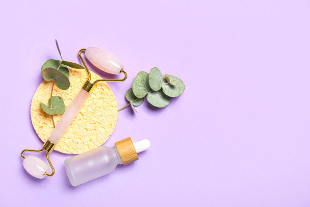 Facial massage tool with cosmetic dropper bottle, sponge and eucalyptus branches on lilac background - 写真・画像