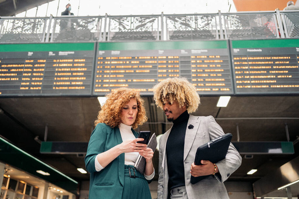 Business woman with formal wear talking with her african coworker and using mobile phone. They are in a train station in Madrid. - Φωτογραφία, εικόνα