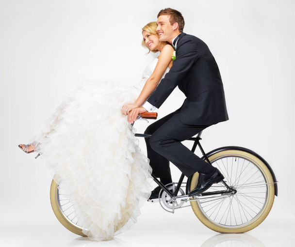 Happy, wedding and couple on a bicycle against a white background, after getting married together. Love, young bride and groom in dress and suit, cycling on a bike in marriage happiness or bliss. - Фото, изображение