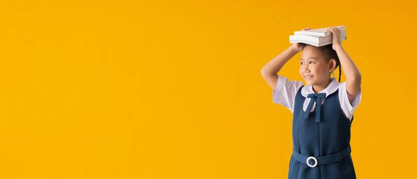 Funny smiling child school girl with glasses hold books on her head, isolated on yellow background with Clipping paths for design work empty free space - 写真・画像