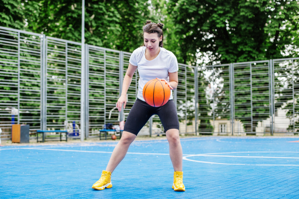 Concept of sports, hobbies and healthy lifestyle. Young athletic woman is training to play basketball on modern outdoor basketball court. - Fotografie, Obrázek