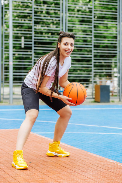 Concept of sports, hobbies and healthy lifestyle. Young athletic woman is training to play basketball on modern outdoor basketball court. - Φωτογραφία, εικόνα